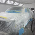 A car covered in blue and yellow plastic wrap is parked on a white concrete floor amidst scattered tools in a well-organized workshop.