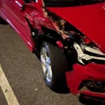 A red car with a broken front wheel, missing tire, and damaged hood and bumper is parked on a street.